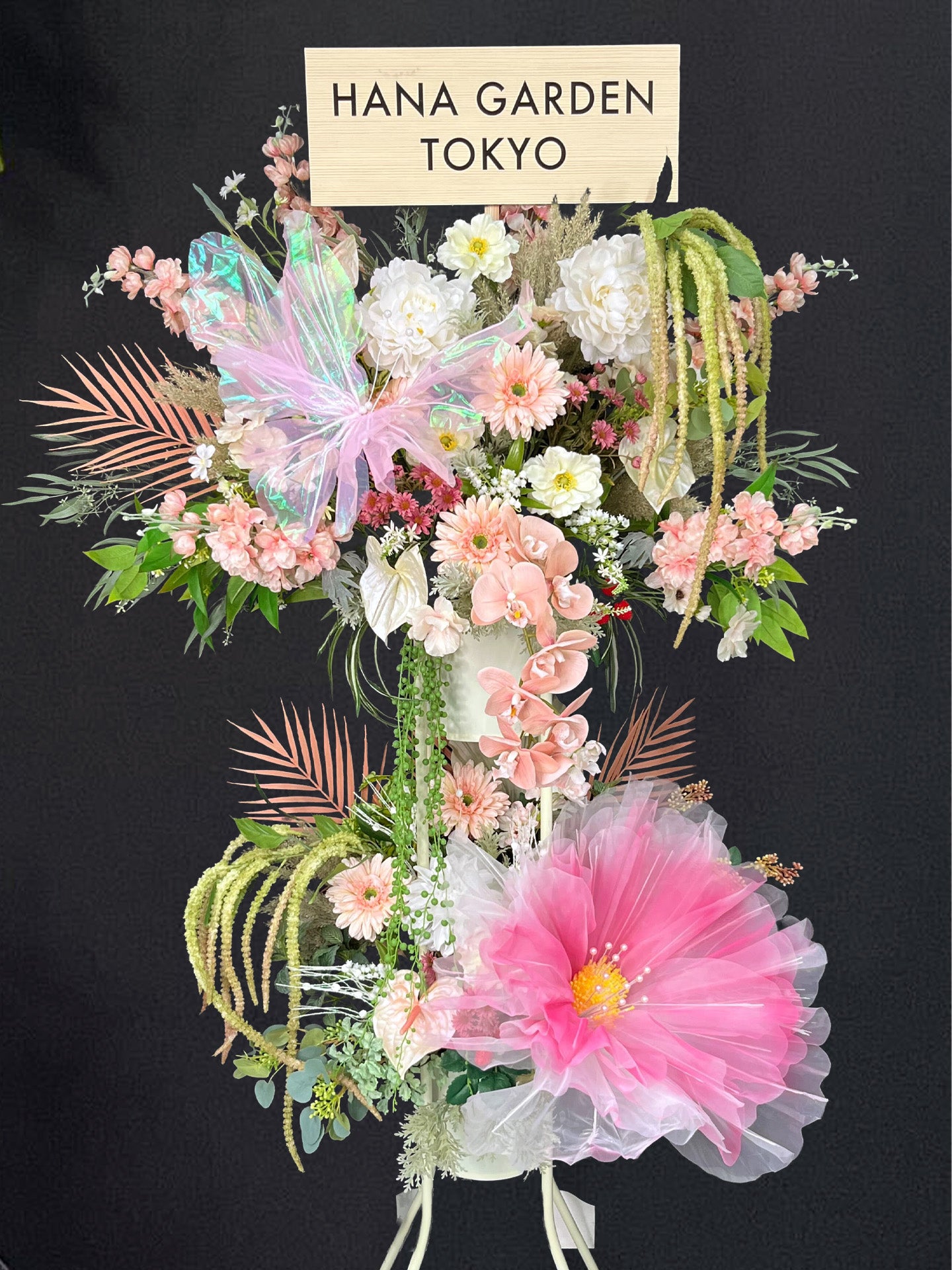 淡いピンクとホワイトの花々にオーロラリボンと蝶が舞う、幻想的で上品なスタンド。
チュールが柔らかく広がるアーティフィシャルフラワーデザインで、公演祝い・イベント装花に最適。