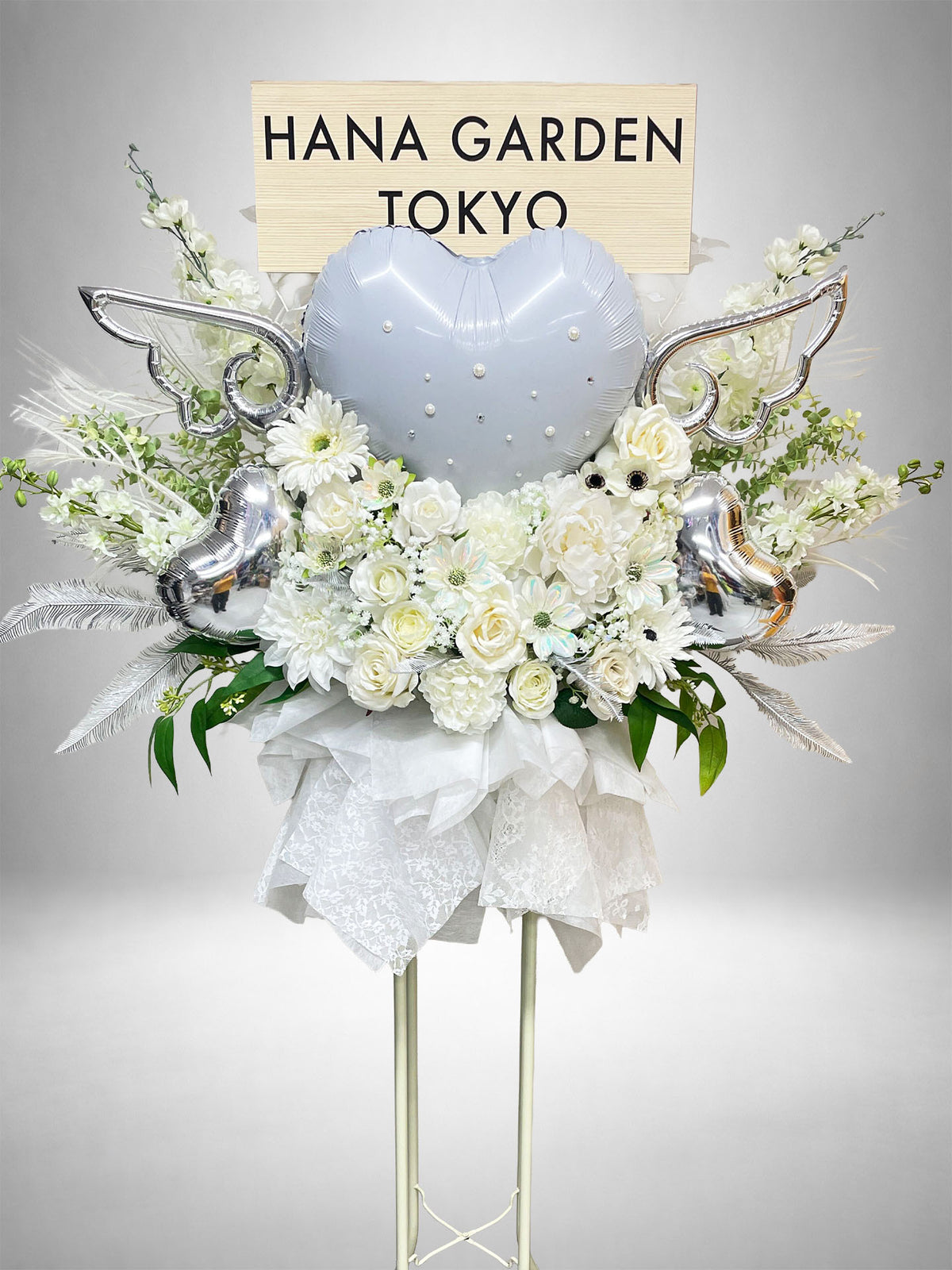 ホワイトの花々にシルバーのハートバルーンと翼を組み合わせた、幻想的で華やかなスタンド花。ライブ・公演祝い、生誕祭、誕生日、イベントなど、ホワイトやシルバーをテーマにした特別なお祝いにおすすめです。