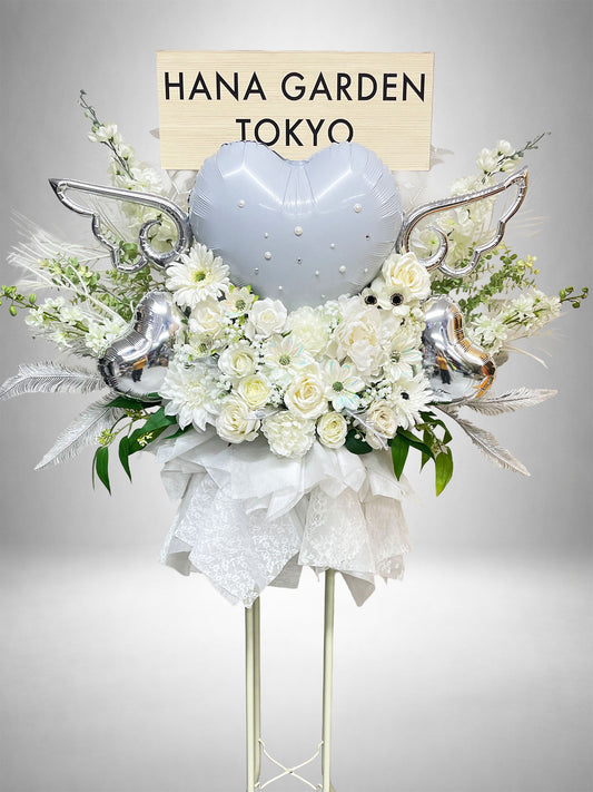 ホワイトの花々にシルバーのハートバルーンと翼を組み合わせた、幻想的で華やかなスタンド花。ライブ・公演祝い、生誕祭、誕生日、イベントなど、ホワイトやシルバーをテーマにした特別なお祝いにおすすめです。