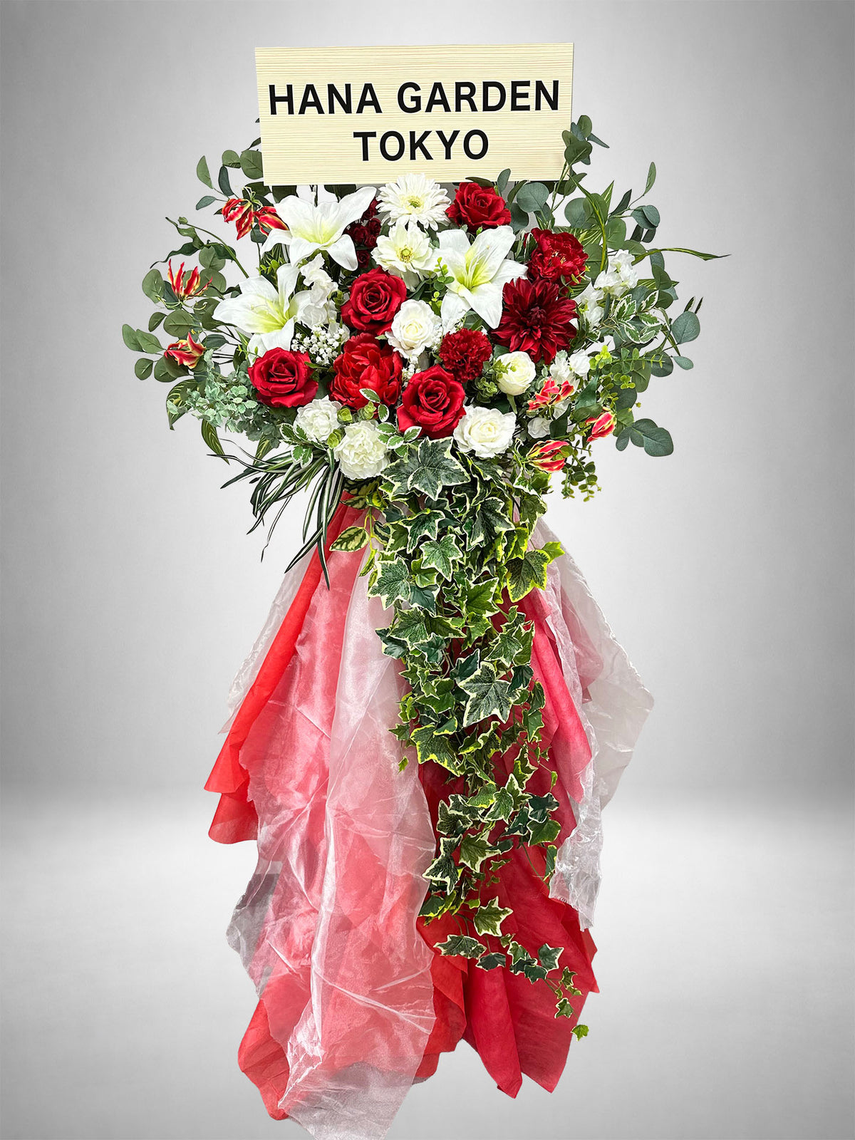 深紅のレッドローズとホワイトの花々に、流れるグリーンを組み合わせたレッドリュミエールスタンド花。ライブ・公演祝い、生誕祭、誕生日、周年祝いなど、華やかで印象的なお祝いにおすすめです。