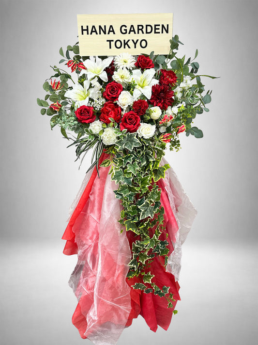 深紅のレッドローズとホワイトの花々に、流れるグリーンを組み合わせたレッドリュミエールスタンド花。ライブ・公演祝い、生誕祭、誕生日、周年祝いなど、華やかで印象的なお祝いにおすすめです。
