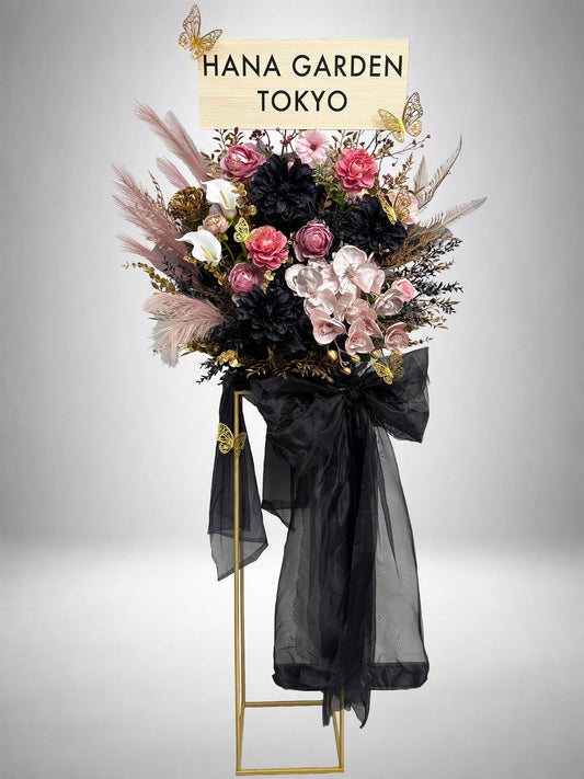 ブラックの大きなリボンに、ピンクの花々とゴールドのアクセントを組み合わせたブラックリュクススタンド花。立体的な花あしらいと洗練されたシルエットが特徴の、ゴージャスで特別感のあるデザインです。ライブ・公演祝い、生誕祭、誕生日、周年祝いなどにおすすめです。
