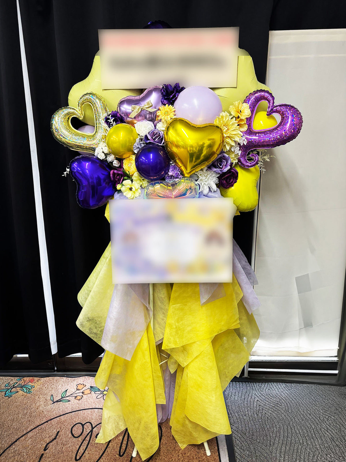 A custom-made flower stand featuring yellow and purple flowers, heart-shaped balloons and yellow fabric decorations.