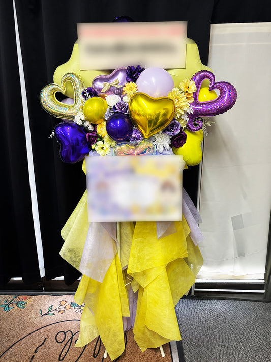 イエローとパープルの花にハート形バルーンとイエローの布装飾を合わせた、オーダーメイドのスタンド花の制作事例です。